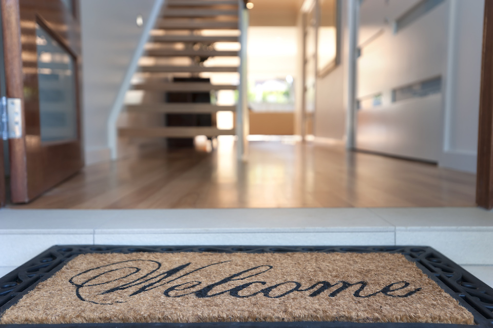 Close up of a welcome mat in front of an inviting house. Focus on foreground